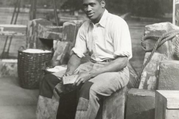 Black and white photo of Paul Robeson, in costume for the 1936 film Showboat, seated on a river wharf near a bale of cotton.
