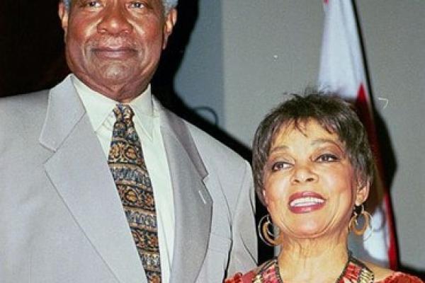 Color photo. Tall, muscular, gray-haired Ossie Davis stands next to petite, radiant Ruby Dee. Davis wears a light gray suit and colorful African Kente tie. Dee wears an elegant red dress with interwoven figures, gold earrings, and a wide African style necklace.