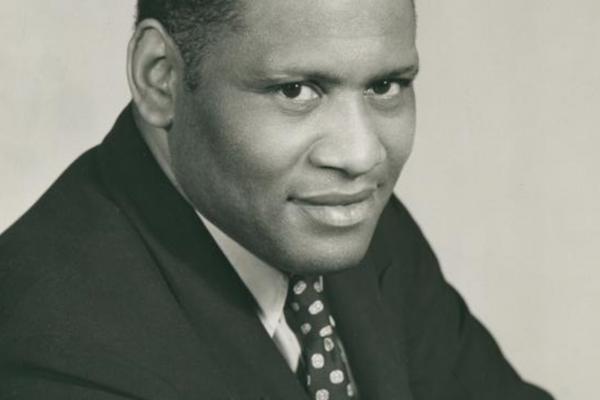 Black and white photo portrait of adult Paul Robeson, wearing a dark suit, white shirt, and white-speckled dark tie.
