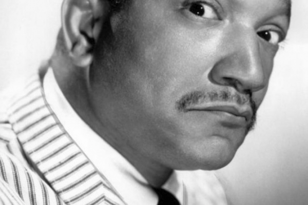 Black and white photo portrait of Redd Foxx in 1966. Foxx has closely cropped hair, a high forehead, and a thin pencil moustache. He is tight-lipped with a playful look on his face, looking askance at the camera. He wears a white jacket, with dark pinstripes, and a dark tie.