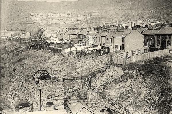 A black and white photo showing a Rhondda Valley, Wales, coal-mining village perched on a hillside in the middle and background of the image. A structure that is part of the colliery is in the foreground.