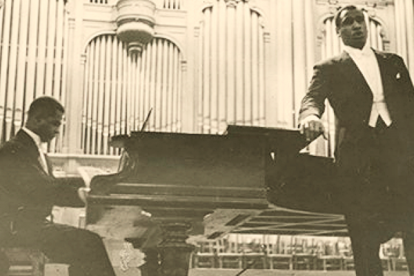 Black and white photo showing Paul Robeson singing on stage in Moscow, 1936, accompanied by pianist Lawrence Brown. Robeson, standing, is wearing a tuxedo; Brown is seated at a grand piano. A large pipe organ forms the background of the image.