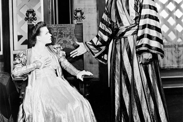 Black and white photo from the Theater Guild’s production of Othello. Paul Robeson’s character Othello is standing at right, with an extended right hand, casting an angry look at Uta Hagen’s seated Desdemona at left. Robeson is dressed in a satin striped robe, and Hagen is wearing a white silk gown.