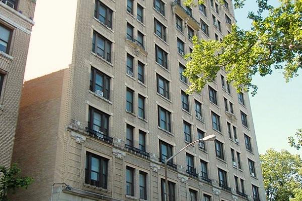 Contemporary color photo of 555 Edgecombe Ave. A multistoried stone building of beige coloring, viewed from the street in late springtime, the season suggested by the bright leafy trees that appear in the image. This otherwise unremarkable building’s only distinctive architectural feature is the Romanesque arch that frames the entrance and includes a small decorative balcony above the arch.