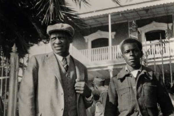Black and white photo of Paul Robeson, shown in the foreground standing beneath a palm tree beside a young African American man who is a volunteer with the International Brigade in the Spanish Civil War. Robeson wears a light-colored suit with vest, striped tie, and English cap. The volunteer wears a dark, buttoned jacket. In the background is a Spanish colonial-style building with a white-wood balcony railing.