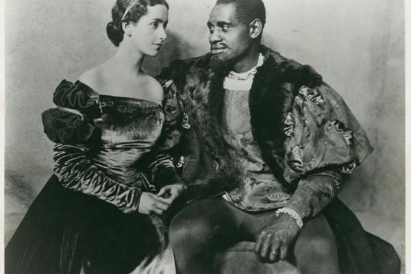 Black and white photo shows Paul Robeson and Peggy Ashcroft seated on stage in period-piece costumes for their roles in the 1930 production of Othello at London’s Savoy Theatre.