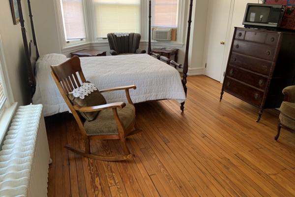 Color photo of Paul Robeson’s bedroom as it was during his final years. The floor is varnished hardwood. The bed, in the background, has dual bedposts above headboard and footboard, with a white coverlet and a single pillow. A white-painted radiator is under the window. A light brown rocking chair with an embroidered throw pillow is between radiator and bed. On the right wall is a dark-brown chest of drawers. A green lounge chair and two small tables are between the bed and three curtained rear windows.