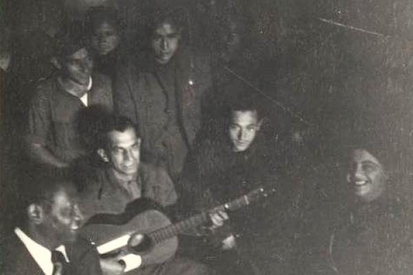 Black and white photo of Paul Robeson singing with freedom fighters at the Madrid battlefront in 1938. Robeson is seated in the lower-left corner of the image beside a man playing a guitar.