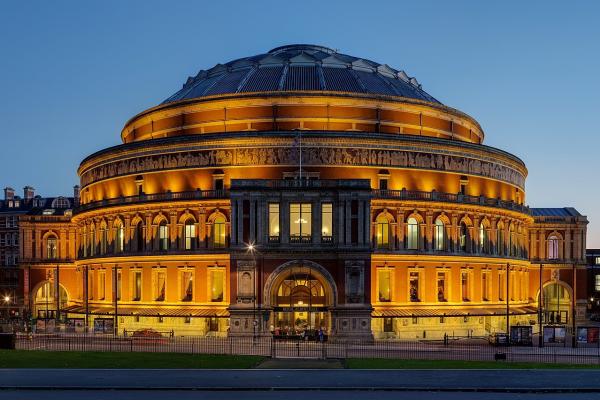 Nighttime color photo of Royal Albert Hall shown in floodlights. A freestanding domed building of iron and glass, designed in an Italianate style, the building’s interior amphitheater is a world-famous concert hall. Red brick dominates the building’s façade.