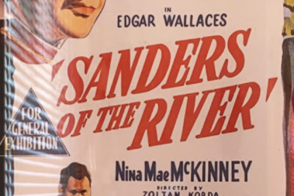 A colorful poster advertises the 1935 film Sanders of the River, showing the film’s stars Paul Robeson and Leslie Banks in their costumes for the film, with bare-chested Robeson posing as a native African chief and Banks wearing a white and brown British pith helmet.