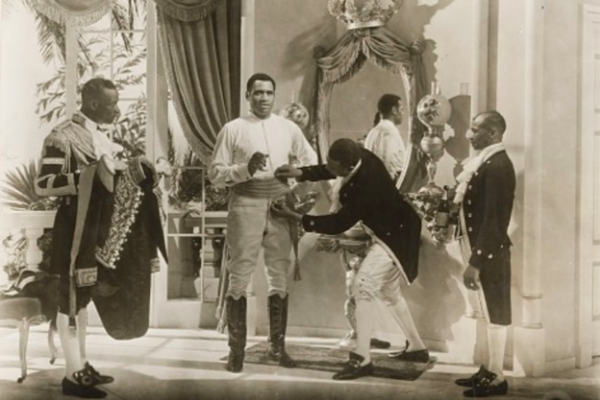 A black and white photo from a scene in the 1933 film Emperor Jones, showing Paul Robeson’s character being dressed in his military uniform by several costumed Black servants.
