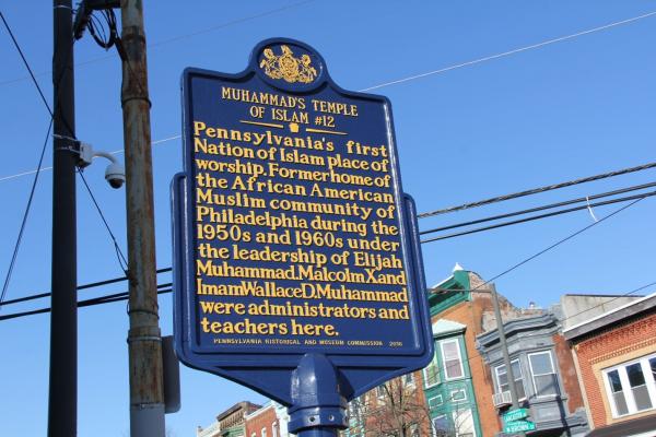 Color photo of a historical marker on a street with modest brick row homes at the intersection of LANCASTER ST and BROWN ST, which reads: MUHAMMAD’S TEMPLE OF ISLAM #12| Pennsylvania’s first Nation of Islam place of worship. Former home of the African American Muslim community of Philadelphia during the 1950s and 1960s under the leadership of Elijah Muhammad. Malcolm X and Imam Wallace D. Muhammad were administrators and teachers here. - Pennsylvania Historical and Museum Commission 2016