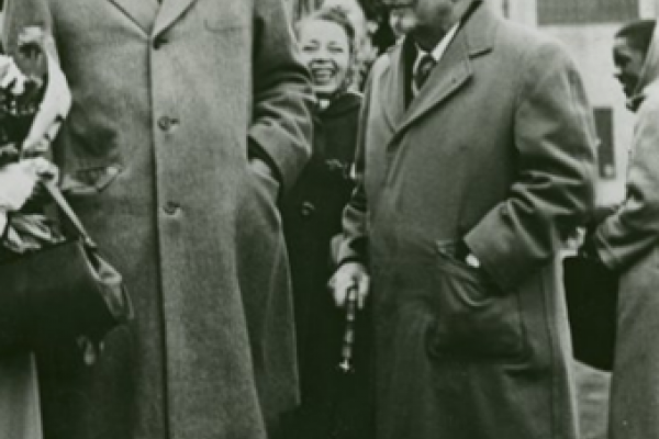 Black and white photo from 1949. Paul Robeson stands in the foreground beside W.E.B. Du Bois. A smiling young woman and what appears to be a small bus appear in the middle ground. Robeson is markedly taller than Du Bois. Both men are wearing gray-toned wool suits and fedoras.